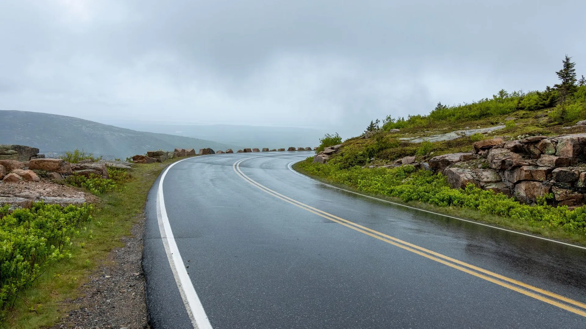 Curving wet road through lush greenery and rocky landscape under cloudy